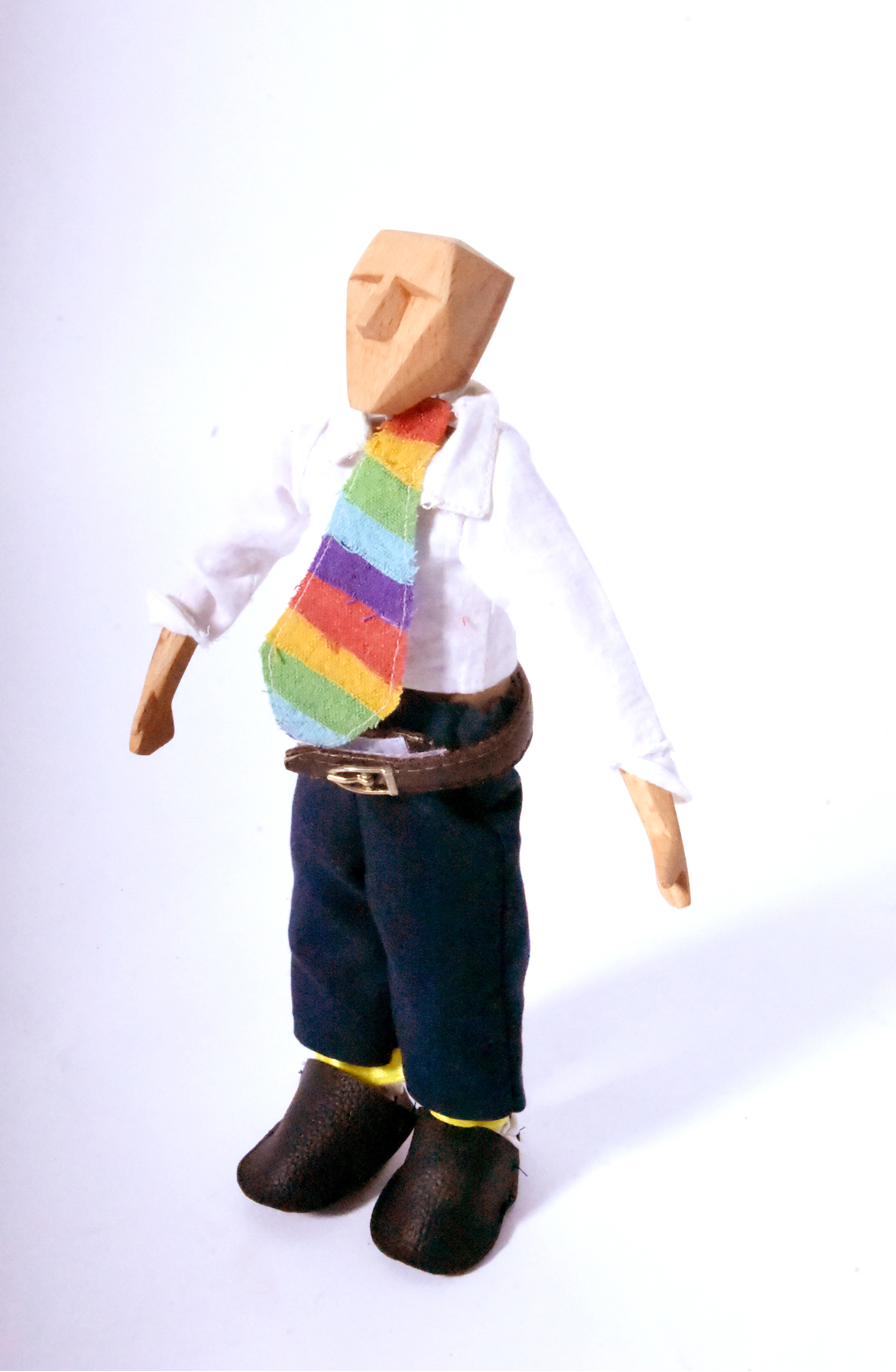 The wooden man dressed in a white shirt, rainbow tie, navy trousers, leather belt, and leather shoes